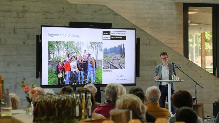 Auf dem Foto ist der Regionalgeschäftsführer des Volksbundes, Jens Effkemann, bei seinem Grußwort zur Ausstellungseröffnung zu erkennen. Er zeigt dem Publikum eine Präsentation u.a. mit dem Bild eines Internationalen Jugendbegegnungsprojektes des Volksbundes in Riga und betont dabei, wie wichtig es sei, neben dem formellen Beitritt ins Deutsche Riga-Komitee, die Mitgliedschaft mit Leben zu füllen und empfiehlt diesbezüglich eine Fahrt mit jungen Leuten in die lettische Hauptstadt. 