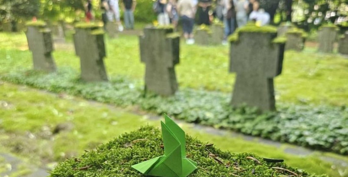 Im Vordergrund ist ein Grabstein mit einer grünen Friedenstaube zu sehen. Im Hintergrund steht eine Gruppe