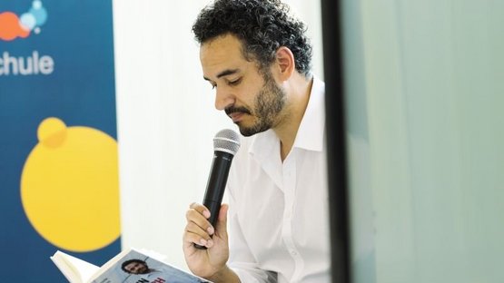 : Im Vordergrund ist der Autor, Dominik Wullers, mit einem strahlend weißen Hemd und einem schwarzen Mikrofon in der Hand zu sehen. Er liest aus seinem geöffneten Buch „Ich bin Deutschland. Plädoyer für einen liberalen Patriotismus“. Im Hintergrund sind etwas unscharf Teile des bunten Logos der VHS Münster zu erkennen.