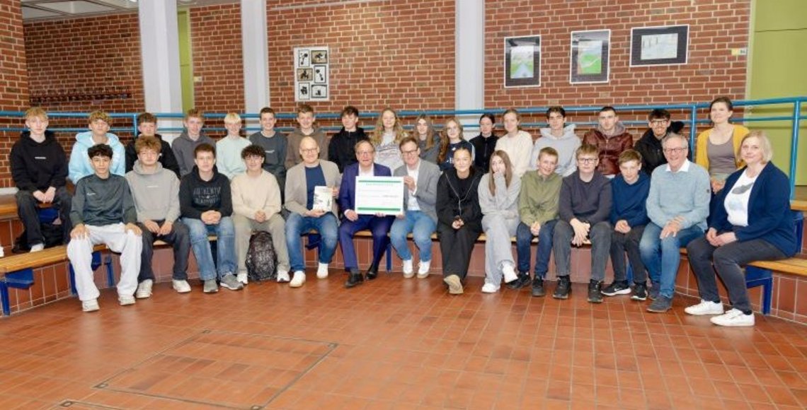 Anlässlich der Übergabe der Spendensammlung wird ein Gruppenfoto der sammelnden Schülerinnen und Schüler der 9b in einem Forum der Schule gemacht. In der Mitte des Fotos sind von links nach rechts der Schulleiter Maarten Willenbrink, der Bürgermeister Dr. Tobias Lehberg und der Regionalgeschäftsführer des Volksbundes, Jens Effkemann, zu erkennen. Der Bürgermeister und der Regionalgeschäftsführer halten den Spendencheck in der Hand. Am rechten Bildrand sind unten von links nach rechts Josef Berkemeier vom Heimatverein sowie Britta Plugge von der Gemeindeverwaltung zu sehen, aber auch in der Reihe darüber die Lehrerin Barbara Skroblin. 