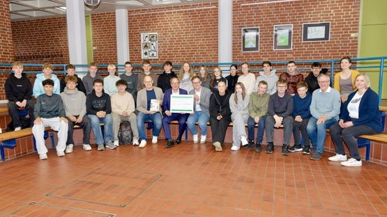Anlässlich der Übergabe der Spendensammlung wird ein Gruppenfoto der sammelnden Schülerinnen und Schüler der 9b in einem Forum der Schule gemacht. In der Mitte des Fotos sind von links nach rechts der Schulleiter Maarten Willenbrink, der Bürgermeister Dr. Tobias Lehberg und der Regionalgeschäftsführer des Volksbundes, Jens Effkemann, zu erkennen. Der Bürgermeister und der Regionalgeschäftsführer halten den Spendencheck in der Hand. Am rechten Bildrand sind unten von links nach rechts Josef Berkemeier vom Heimatverein sowie Britta Plugge von der Gemeindeverwaltung zu sehen, aber auch in der Reihe darüber die Lehrerin Barbara Skroblin. 
