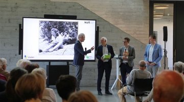 Über die Köpfe des Publikums hinweg ist links der Kreisdirektor des Kreises Borken, Dr. Ansgar Hörster zu sehen, der angesichts der doppelten Ausstellungseröffnung im kult Westmünsterland zwei Präsente an Paul Glaser (Initiator der Ausstellung „Die Tänzerin von Auschwitz“) und Jens Effkemann (Regionalgeschäftsführer des Volksbundes) verteilt. Daneben steht rechts der Bürgermeister der Stadt Vreden, Tom Tenostendarp. Im Hintergrund wird ein großes Foto der lachenden Tänzerin Roosje Glaser am Strand gezeigt. 