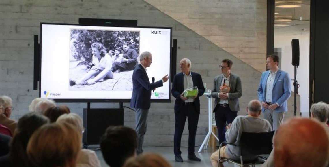 Über die Köpfe des Publikums hinweg ist links der Kreisdirektor des Kreises Borken, Dr. Ansgar Hörster zu sehen, der angesichts der doppelten Ausstellungseröffnung im kult Westmünsterland zwei Präsente an Paul Glaser (Initiator der Ausstellung „Die Tänzerin von Auschwitz“) und Jens Effkemann (Regionalgeschäftsführer des Volksbundes) verteilt. Daneben steht rechts der Bürgermeister der Stadt Vreden, Tom Tenostendarp. Im Hintergrund wird ein großes Foto der lachenden Tänzerin Roosje Glaser am Strand gezeigt. 