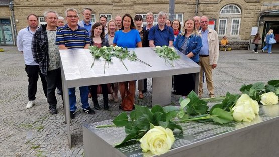 Auf dem Bild ist die Gruppe hinter einer von zwei Gedenkstelen vor dem Hauptbahnhof in Bielefeld zu sehen. In der Mitte ist die Regierungspräsidentin Anna Katharina Bölling mit blauem Kleid zu erkennen. Zur Erinnerung an die deportierten Menschen, die auf den Stelen aufgeführt sind, hat die Gruppe zuvor weiße Rosen darauf niedergelegt.